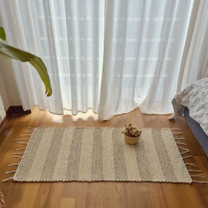 Può includere: Un tappeto rettangolare a righe verticali beige e grigie, con frange sui lati corti. Il tappeto è posizionato su un pavimento in legno, con un piccolo cestino intrecciato con fiori secchi sopra. Tende bianche trasparenti sullo sfondo.