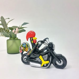 Peut inclure: Une figurine de moto en céramique noire avec un coq coloré qui la chevauche. Le coq a une crête et une barbe rouges, et ses plumes sont peintes de différentes couleurs, dont le jaune, le vert et le bleu.