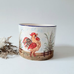Könnte beinhalten: Ein weißer Keramiktopf mit blauem Rand zeigt ein handgemaltes Design eines Hahns mit roten, gelben und braunen Federn, der auf einer grünen Wiese steht.