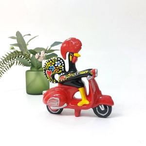 Puede incluir: Una figura de gallo de cerámica roja y negra montando un scooter rojo. El gallo tiene un diseño pintado de colores en sus plumas de la cola.