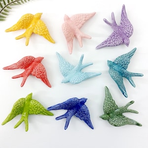 Op de afbeelding: Een verzameling van negen keramische zwaluwbeeldjes in verschillende kleuren, waaronder geel, roze, paars, rood, lichtblauw, turquoise, groen en donkerblauw. Elke vogel heeft gedetailleerde vleugel- en staartveertexturen.