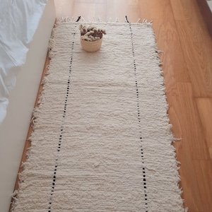Puede incluir: Una alfombra tejida blanca con costuras negras y bordes deshilachados. La alfombra está colocada sobre un suelo de madera.