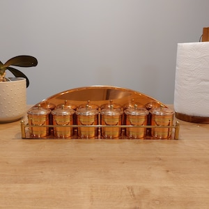 May include: A set of eight copper spice jars with brass lids and a copper rack. The jars are arranged in a row on the rack, which is shaped like a half-circle.