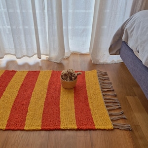 Puede incluir: Una alfombra rectangular con rayas verticales naranjas y amarillas. La alfombra tiene un borde con flecos y está colocada sobre un suelo de madera. Una pequeña cesta tejida llena de flores secas está sobre la alfombra.