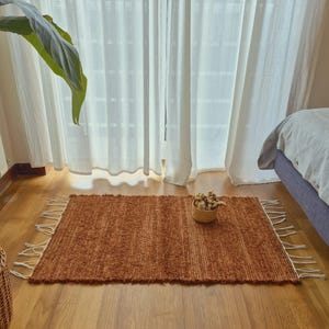 Puede incluir: Una alfombra rectangular de color óxido con una superficie texturizada y bordes con flecos blancos. La alfombra está colocada sobre un suelo de madera frente a una ventana con cortinas blancas. Una pequeña cesta tejida con flores secas está sobre la alfombra.