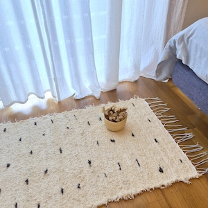 May include: Cream-colored rectangular rug with black accents and fringe. A small woven basket filled with dried flowers sits on the rug. The rug is placed on a wooden floor, near a bed and white curtains.