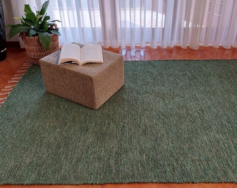 May include: A green woven rug with a light brown ottoman and a book on top. The rug has a fringed edge.