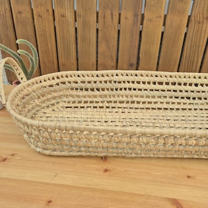 May include: A woven, oval-shaped basket with a natural, light tan color. The basket has a textured surface and is made of woven material. The basket is on a wooden surface with a wooden background.