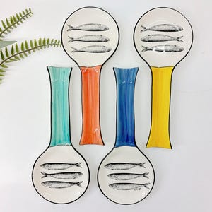 Può includere: Quattro poggiacucchiaio in ceramica su sfondo bianco. Ogni poggiacucchiaio ha un manico di colore diverso: verde acqua, arancione, blu e giallo. I poggiacucchiaio hanno un bordo nero e presentano un'illustrazione in bianco e nero di tre pesci.