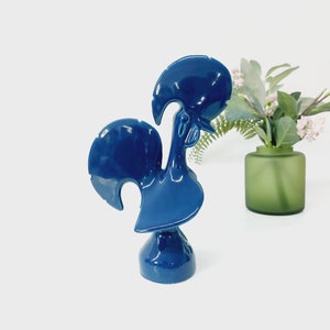 Puede incluir: Una figurita de gallo de cerámica azul con una cola levantada y una cabeza detallada. El gallo está de pie sobre una base.