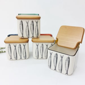 Puede incluir: Un juego de cuatro recipientes de cerámica con tapas de madera. Los recipientes son blancos con un patrón repetido de peces en blanco y negro. Las tapas son rectangulares con un acabado de madera natural y detalles de color.