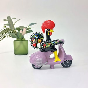Peut inclure: Figurine en céramique d'un coq noir avec une crête rouge et des bottes jaunes, conduisant un scooter lavande. La queue du coq est ornée de motifs floraux colorés. Le scooter a des roues noires.