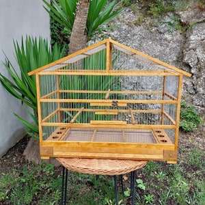 Puede incluir: Una jaula de pájaros de madera con techo a dos aguas y laterales de malla metálica. La jaula tiene una base rectangular con un suelo de malla metálica y pequeñas aberturas circulares. Está hecha de madera clara y está sobre una mesa de mimbre.
