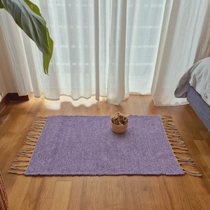 May include: A rectangular, lavender-colored rug with a woven texture and fringed edges. A small, woven basket filled with dried flowers sits on the rug. The rug is placed on a hardwood floor near a window with white curtains.