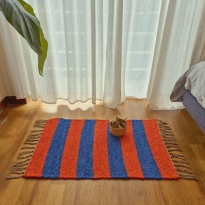 Peut inclure: Tapis rectangulaire à rayures verticales orange et bleu. Le tapis est bordé de franges et posé sur un parquet. Un petit bol en osier contenant des fleurs séchées est posé sur le tapis.