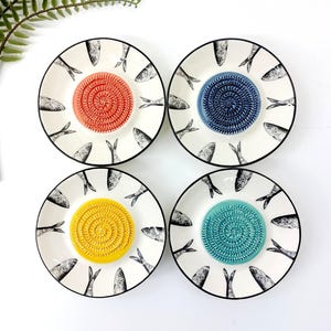 Peut inclure: Quatre assiettes rondes en céramique ornées d'un motif de poissons noir et blanc sur le bord. Chaque assiette possède un centre texturé en forme de spirale, de couleur unie différente : orange, bleu foncé, jaune et bleu sarcelle, pour râper.