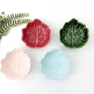 Op de afbeelding: Vier blad-vormige keramische schalen in rood, groen, lichtblauw en roze. Elke schaal heeft een gedetailleerd bladaderontwerp. De schalen zijn gerangschikt op een wit oppervlak, met een takje groen.