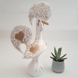 May include: White ceramic rooster figurine with gold painted details. The rooster has a large, ornate crest and a heart-shaped design on its chest.