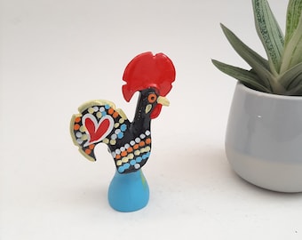 May include: A colorful ceramic rooster figurine with a red comb and black, white, and yellow painted details. The rooster is standing on a blue base.