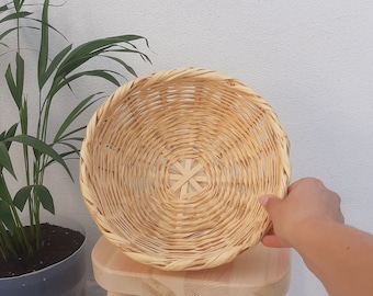 May include: A round, woven basket made of natural materials. The basket is light brown and has a circular shape. It is sitting on a wooden stool.
