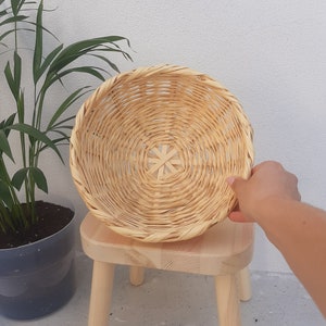 Op de afbeelding: Een ronde, geweven mand gemaakt van natuurlijke materialen. De mand is lichtbruin en heeft een ronde vorm. Hij staat op een houten kruk.