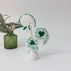 Puede incluir: Figurita de gallo de cerámica blanca con detalles pintados en verde. El gallo tiene una gran cresta verde y una cola verde con un diseño de corazón.