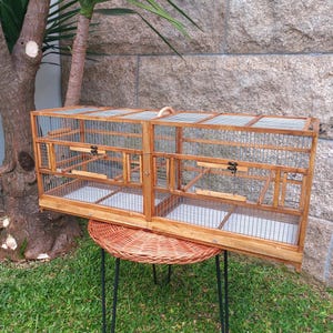 Puede incluir: Una jaula de pájaros de madera con forma rectangular, con malla de alambre y un asa en la parte superior. La jaula tiene dos compartimentos, cada uno con una pequeña puerta y una bandeja en la parte inferior. Está sobre una mesa redonda tejida.