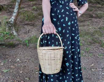 May include: A woman in a black dress with a blue floral pattern is holding a woven wicker basket with a handle.