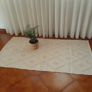 May include: A white woven rug with a diamond pattern. The rug is on a wooden floor and a potted plant is in the foreground.