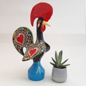 Puede incluir: Una figura de gallo negro y rojo con detalles pintados de colores. El gallo está de pie sobre una base azul y tiene una cresta y una barbilla rojas.