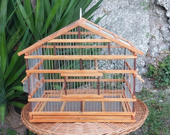 May include: A wooden birdcage with a wire mesh interior. The cage has a peaked roof and two small side doors. The cage is sitting on a wicker table.