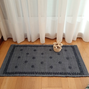 Könnte beinhalten: Ein dunkelblauer gewebter Teppich mit geometrischem Muster. Der Teppich hat einen weißen Rand und liegt auf einem Holzboden. Ein kleiner Korb mit getrockneten Blumen steht auf dem Teppich.