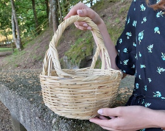May include: A round, light brown woven basket with a handle. The basket is made of thin, natural-colored reeds. The handle is arched and attached to the sides of the basket. The basket is held by a person in a black floral dress.