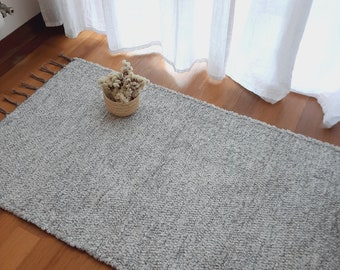 Puede incluir: Una alfombra gris y esponjosa con una textura sutil. La alfombra está colocada sobre un suelo de madera con luz natural que entra por una ventana.