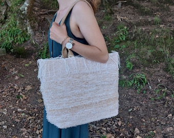 May include: A white woven tote bag with fringe detailing. The bag has a brown leather strap and is being held by a person wearing a teal dress.