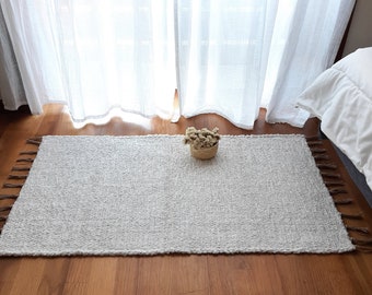 Puede incluir: Una alfombra blanca tejida con flecos en los bordes. La alfombra está sobre un suelo de madera delante de una cortina blanca transparente.