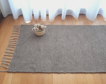 Puede incluir: Una alfombra gris tejida con flecos en los bordes. La alfombra está sobre un suelo de madera y hay una pequeña cesta con flores secas encima de la alfombra.