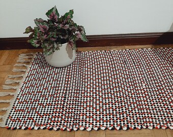 May include: A black and white checkered rug with red accents. The rug has a fringe border and is made of a woven material. A potted plant is sitting on the rug.