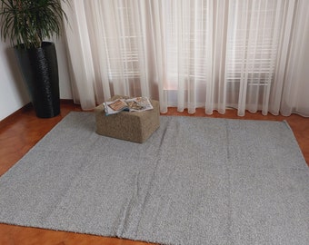 May include: A gray shag rug with a light gray ottoman in the center. The ottoman is topped with a stack of magazines. The rug is in front of a window with white sheer curtains.