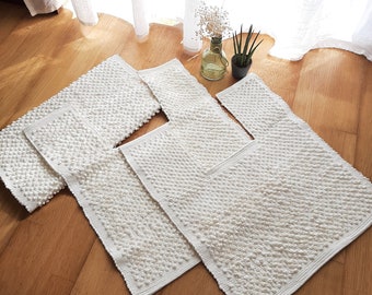 Op de afbeelding: Drie witte badmatten met een textuur van verhoogde stippen. De matten zijn in een verspringend patroon op een houten vloer gelegd.