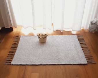 Puede incluir: Una alfombra blanca tejida con flecos en los bordes, colocada sobre un suelo de madera delante de una cortina blanca transparente. Una pequeña cesta de flores secas se encuentra sobre la alfombra.