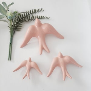 May include: Three pink ceramic bird wall decorations in different sizes. The birds are in a flying position with their wings spread out.