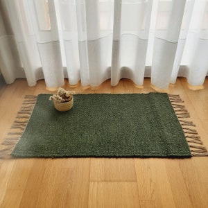 May include: A dark green woven rug with fringed edges. The rug is on a wooden floor and a small basket with dried flowers is in the center of the rug.