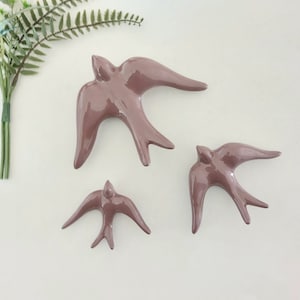 Pode incluir: Três andorinhas de cerâmica em cor rosa poeirento para decoração de parede. As andorinhas estão em voo e são de tamanhos diferentes.