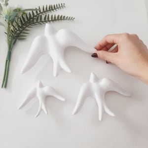 Op de afbeelding: Drie witte keramische vogelwanddecoraties in verschillende maten. De vogels bevinden zich in een vliegende pose met uitgespreide vleugels.