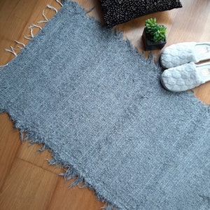 May include: A gray, fluffy rug with frayed edges. The rug is laid out on a wooden floor.