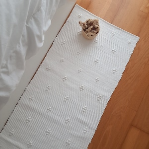 Puede incluir: Alfombra blanca tejida con un patrón de flores texturizado y en relieve. La alfombra está colocada sobre un suelo de madera dura.