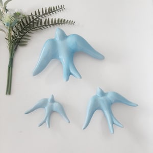 Op de afbeelding: Drie lichtblauwe keramische zwaluwen met uitgespreide vleugels zijn gerangschikt op een wit oppervlak. De zwaluwen variëren in grootte, met de grootste bovenaan en de kleinste onderaan. Een takje groene en witte kunstbloemen staat in de linkerbovenhoek.