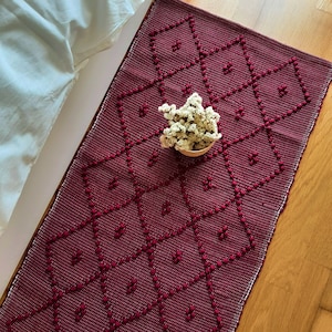 May include: A burgundy woven runner with a diamond pattern and raised dot accents. A small woven basket holds dried white flowers. The runner is placed on a wooden surface, with a white sheet visible in the background.