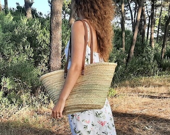May include: A woman wearing a white floral maxi dress with a side slit carries a large woven basket bag with leather straps in a wooded area.